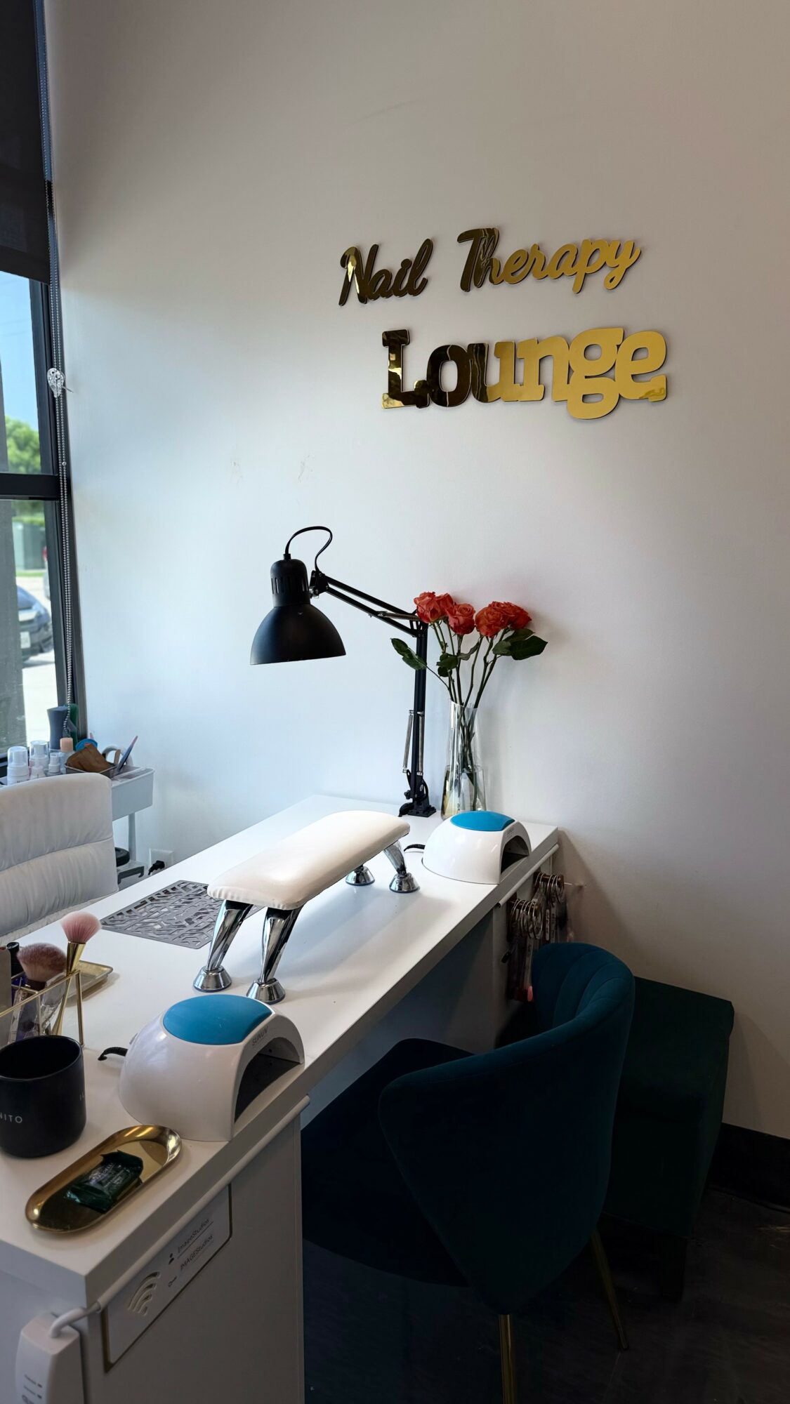 Nail therapy lounge sign on wall, manicure table with lamp, flowers, and chairs in a bright room.