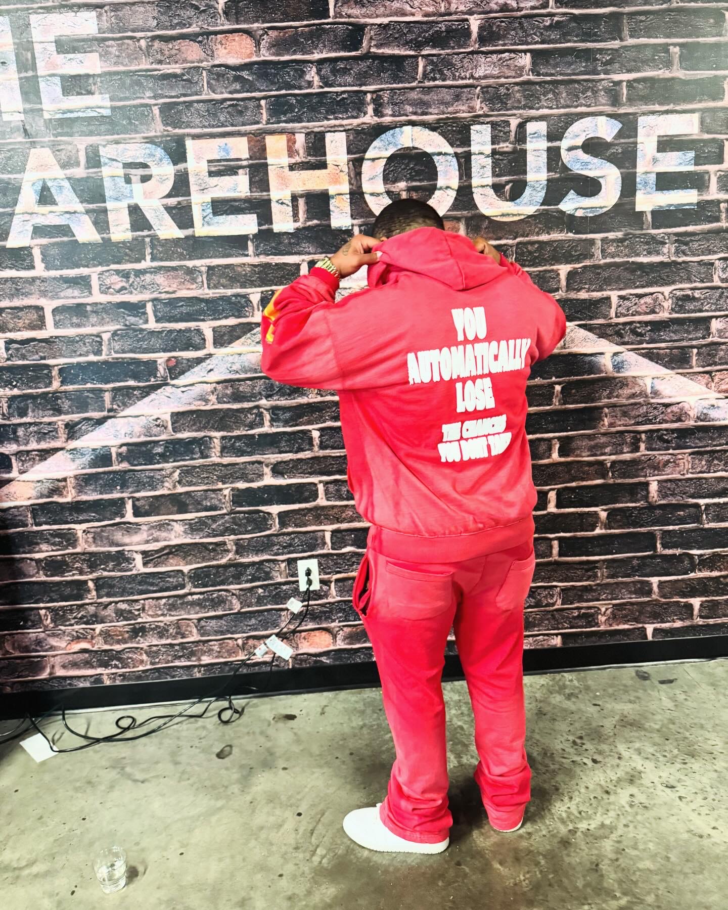 Person in red jacket and pants facing a brick wall with partially visible white text, spray painting the wall, on a concrete floor.