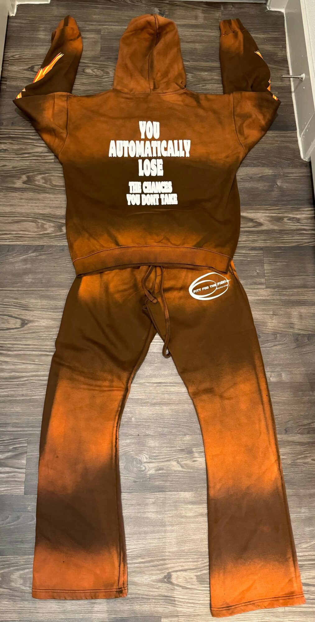 Brown hoodie and pants laid flat on a wooden floor, with text on the hoodie and a logo on the pants.
