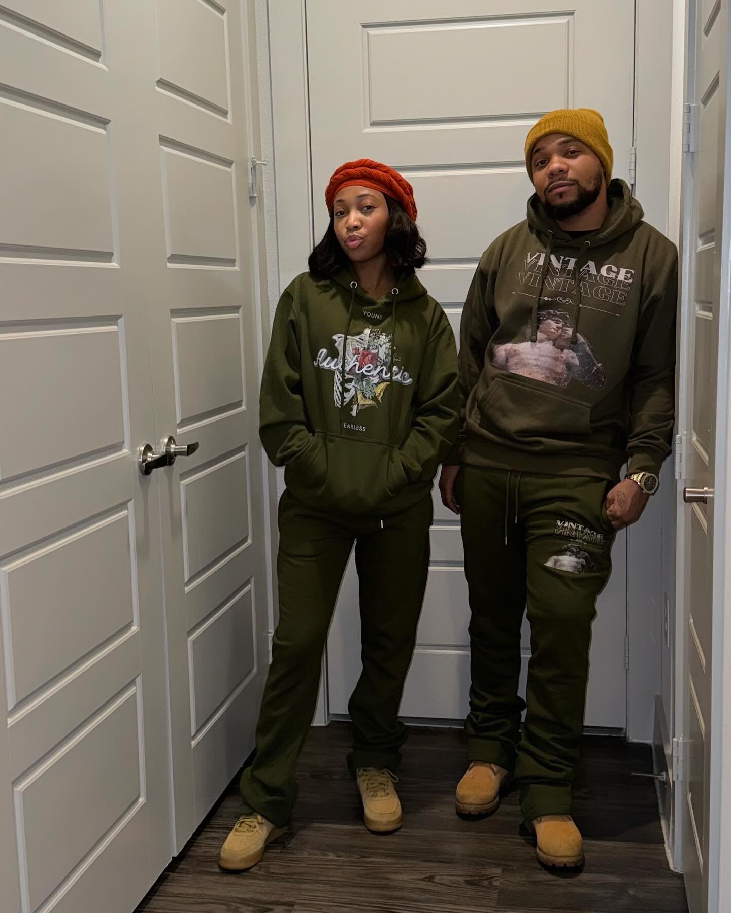 Two people standing in a narrow hallway with white paneled doors, wearing casual clothing and hats, looking at the camera.