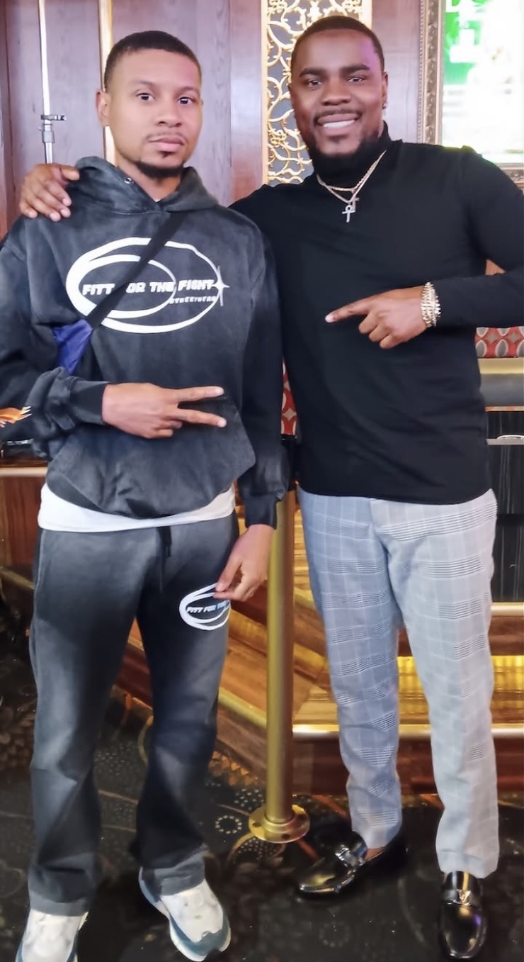 Two men standing indoors, one with arm around the other, both smiling, one making a peace sign, casual clothing, background with patterned wall.