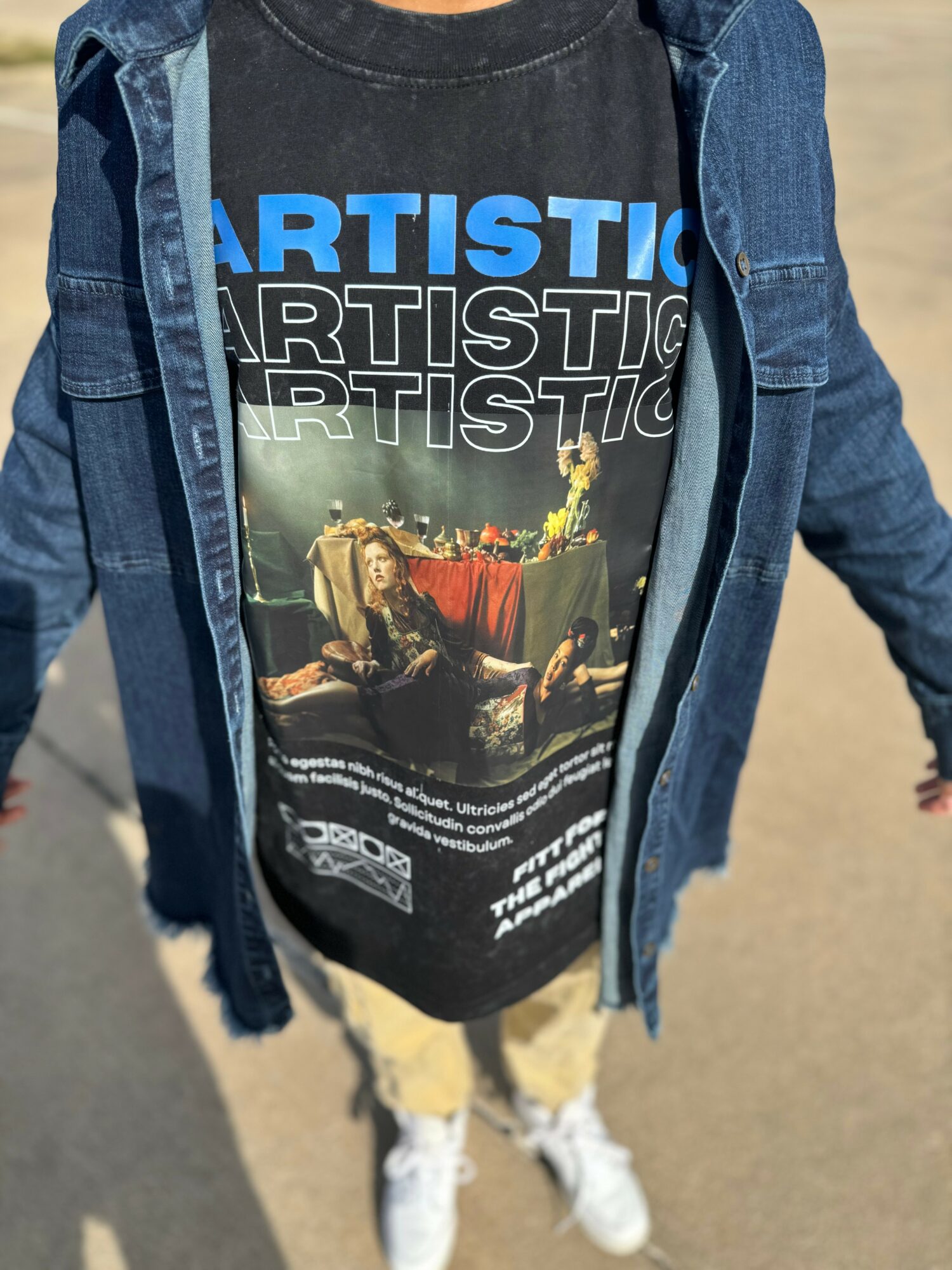 Person wearing a black graphic T-shirt with the word ARTISTIC and an image of a person sitting at a table, layered with a blue denim jacket.