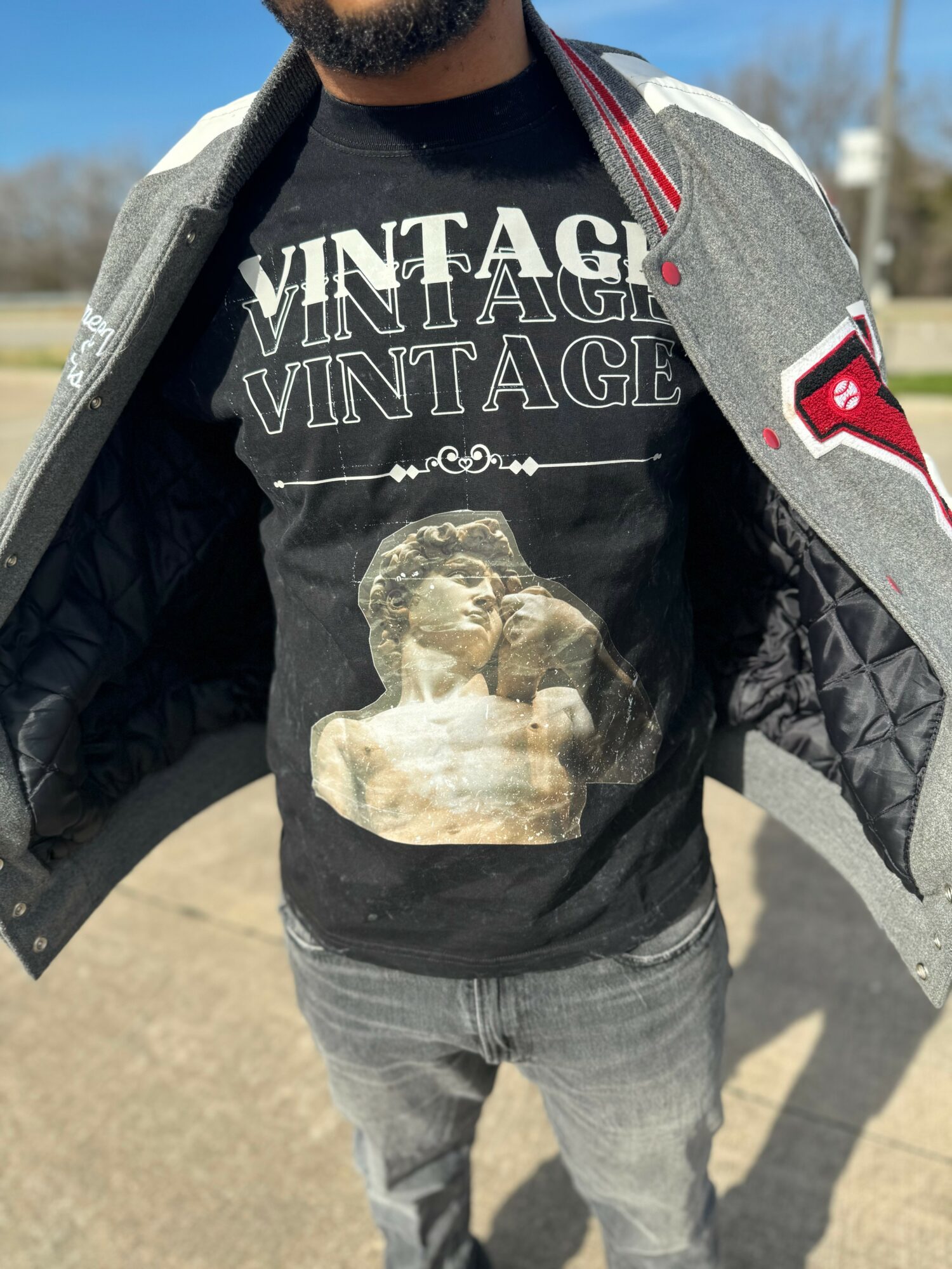Person wearing a black T-shirt with vintage-style graphic, gray jacket, and jeans, standing outdoors with a clear sky.