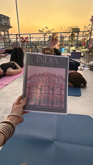Person holding a book titled 'India' with an illustration of a historic building, on a rooftop during sunset, with yoga mats and people in the background.