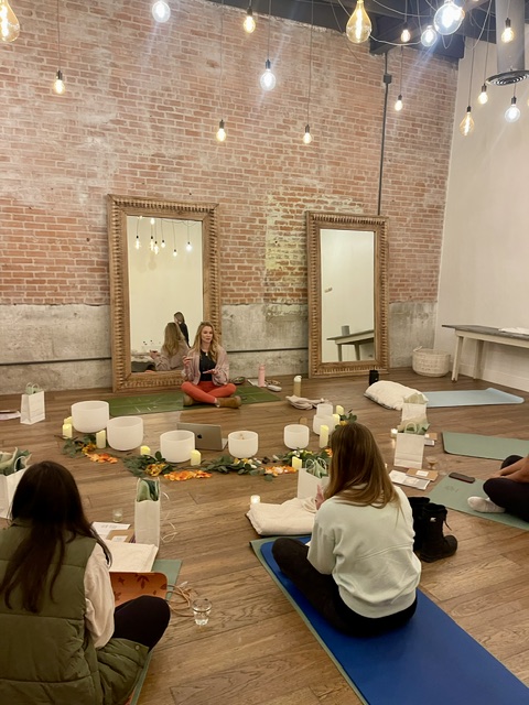 People sitting on yoga mats in a room with brick wall, mirrors, and hanging lights, participating in a yoga or meditation session.