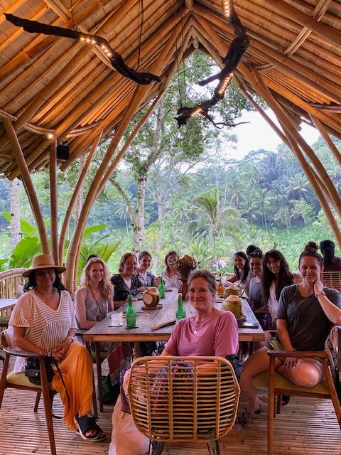 Group of people sitting and standing around a table in a bamboo structure with greenery outside, some smiling and talking.