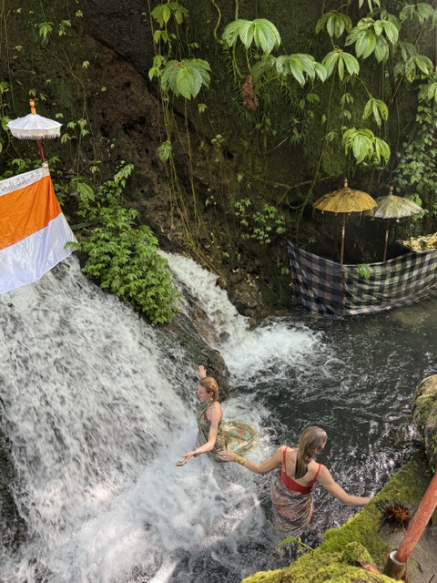 Two children play in a small waterfall surrounded by green plants, with one child leaning over the water.