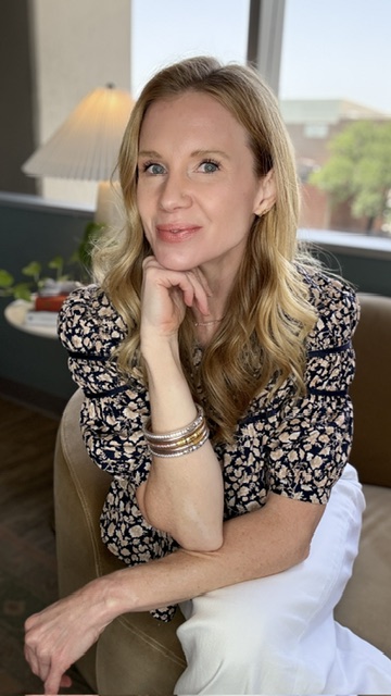 Woman with blonde hair sitting indoors near a window, resting her chin on her hand, smiling gently.