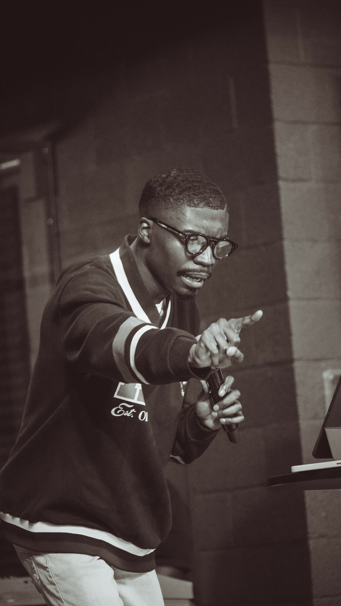 Young man with glasses pointing at a screen, indoors, in black and white.