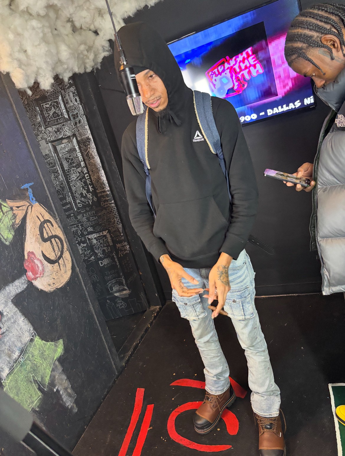 Person wearing a black hoodie, jeans, and brown boots standing indoors near a wall with graffiti art, holding a phone.