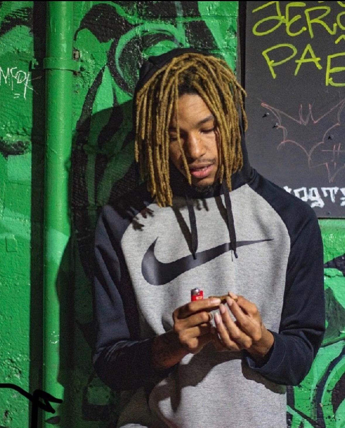 Young man with dreadlocks looking at a lighter, standing against a green graffiti wall.