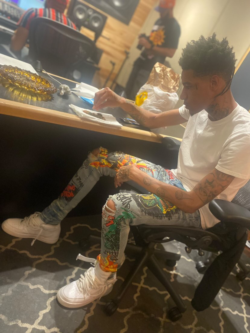 Young man with tattoos sitting at a table, wearing colorful pants and white sneakers, in a room with people and equipment.