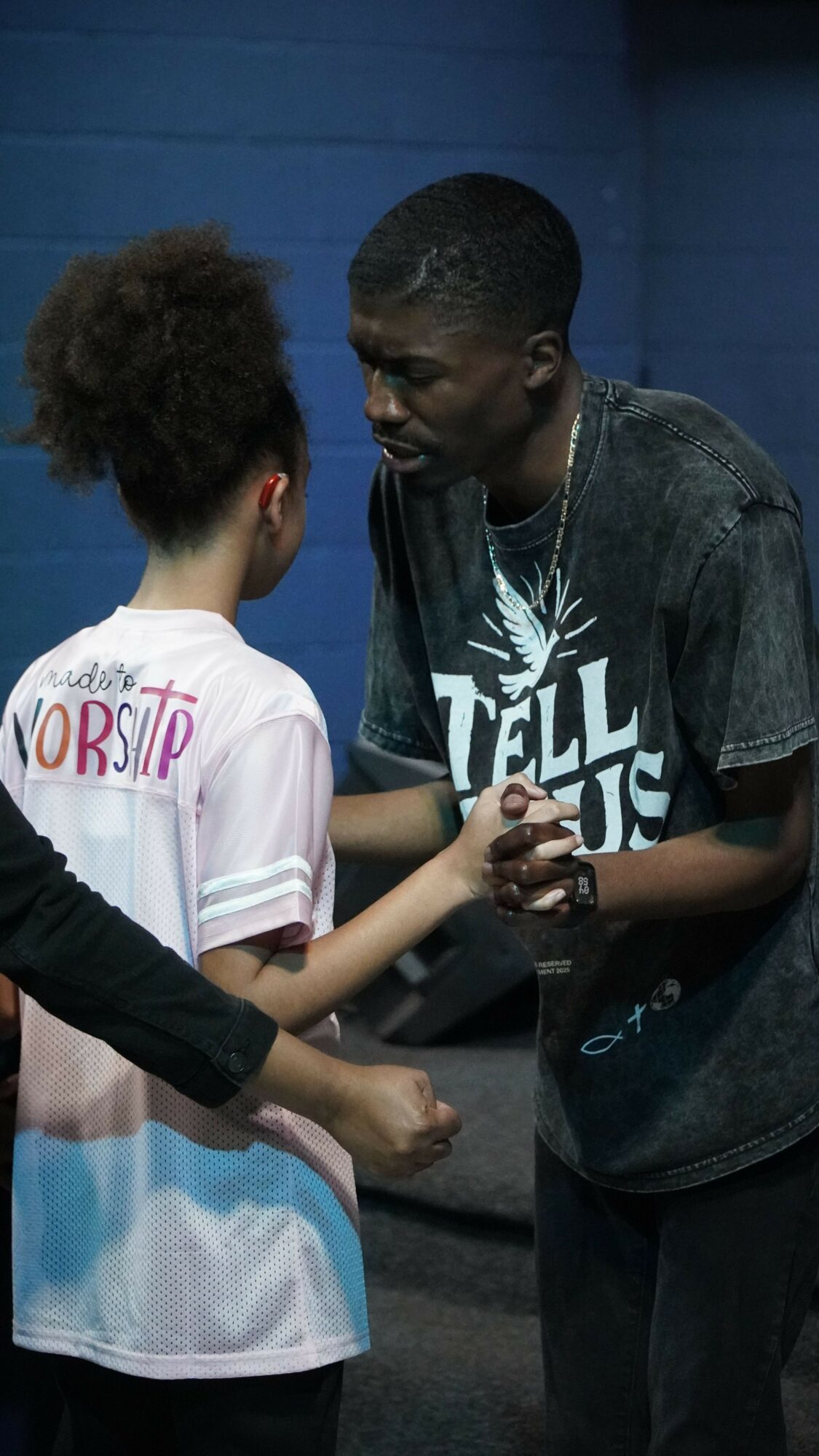Two young people standing and talking indoors, one with curly hair and the other with short hair, against a blue wall.