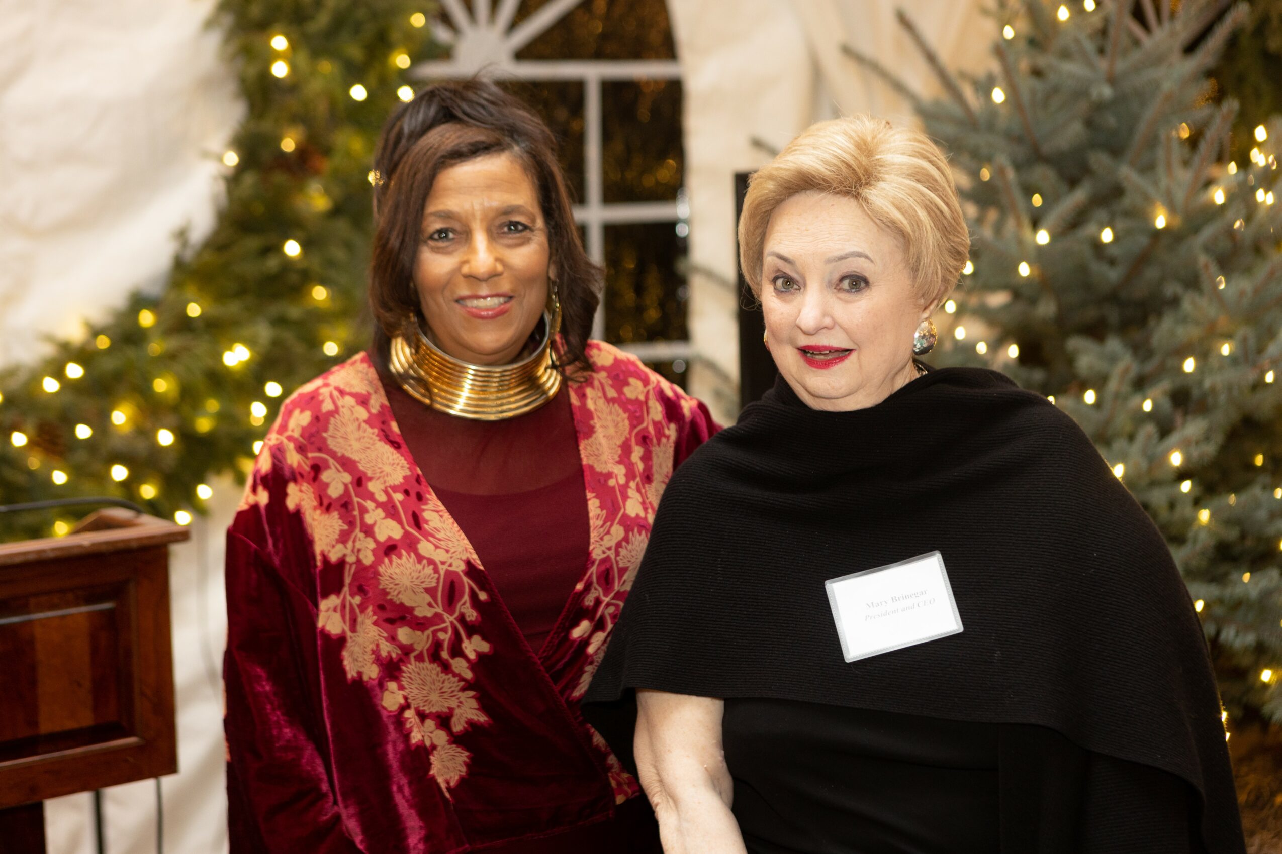 Two women standing together in front of decorated Christmas trees, smiling, with festive lights in the background.