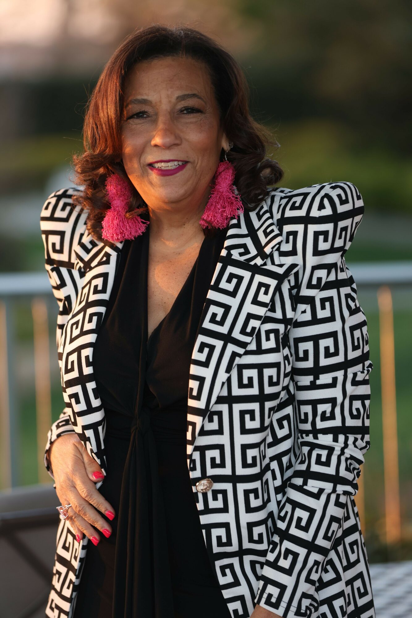 Woman with red hair and pink earrings smiling outdoors, wearing a black and white patterned jacket.