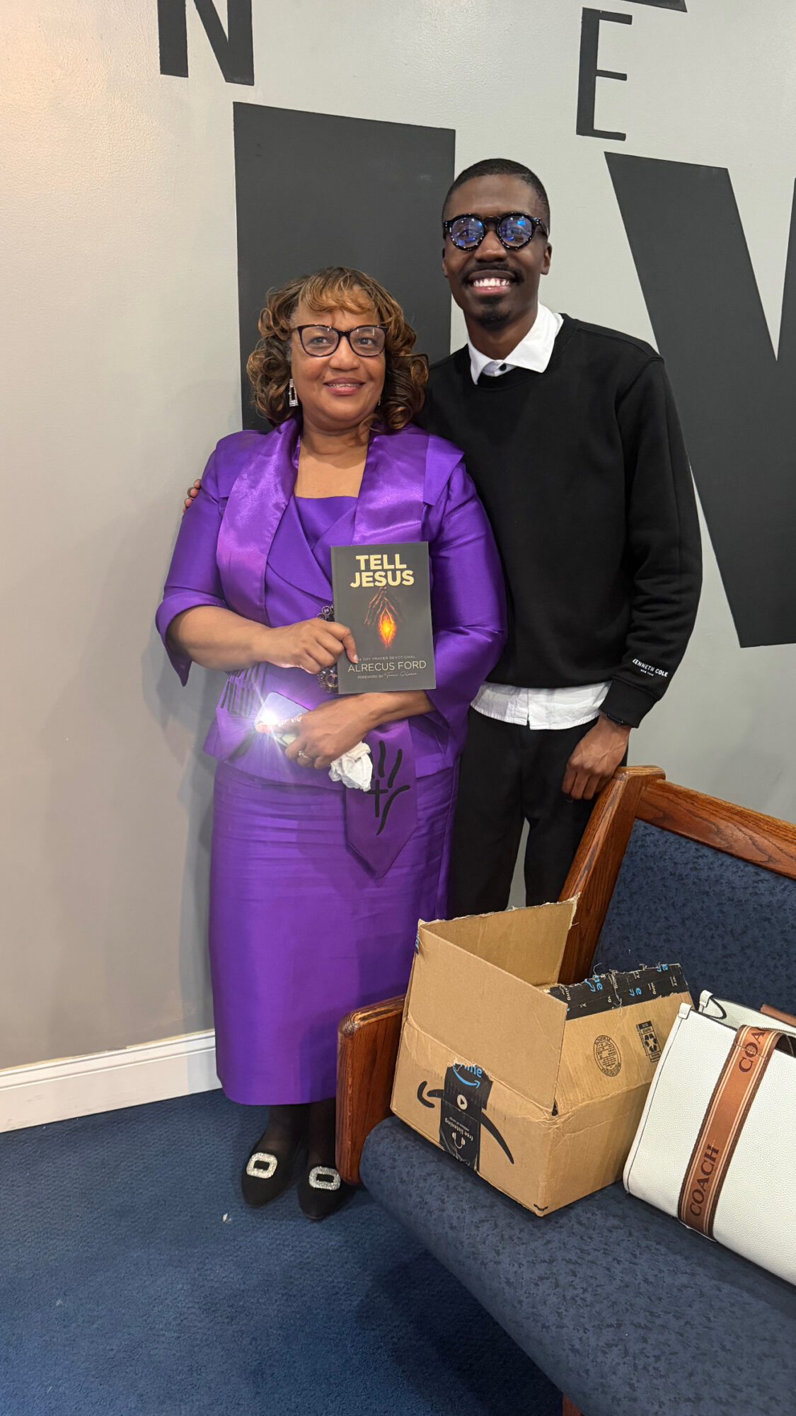 A woman and a man stand together indoors, smiling, with a book and a box on a nearby chair.