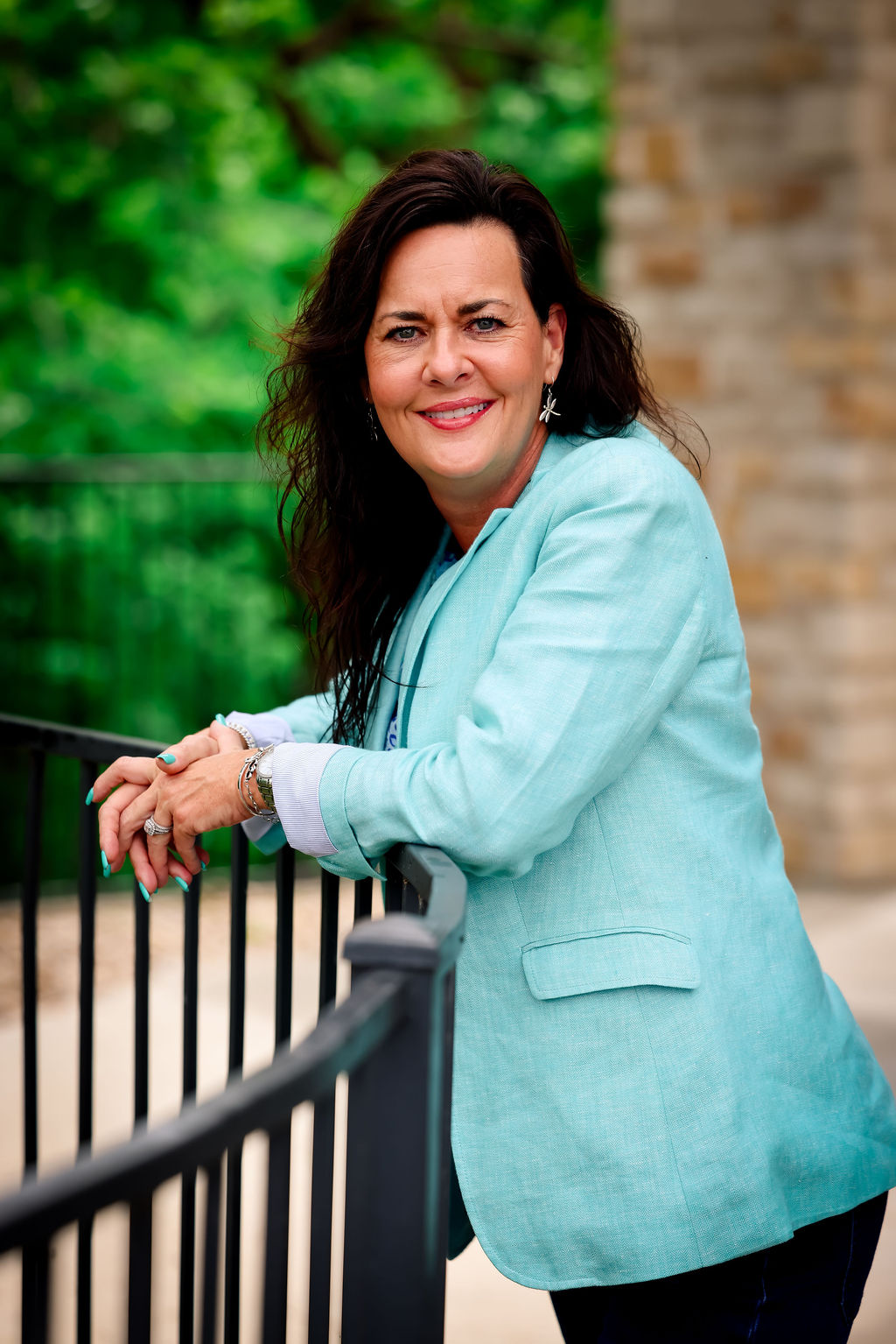 Woman with dark hair smiling outdoors, wearing a light blue blazer, standing near a black fence with greenery and a brick wall in background.