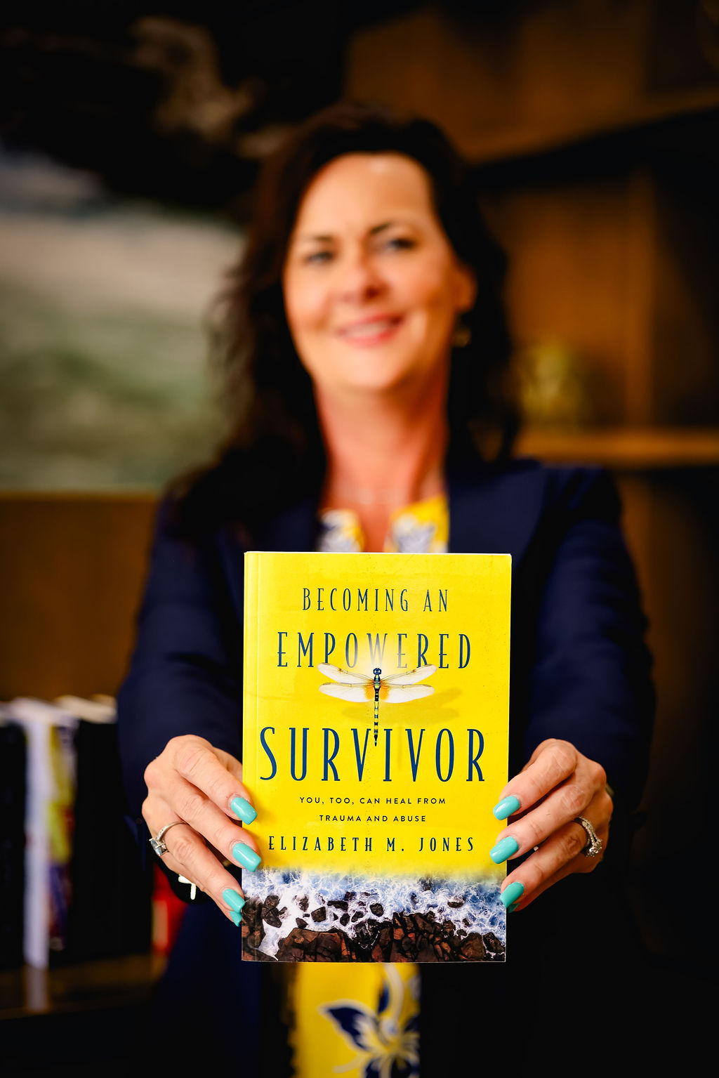 Woman holding a book titled 'Becoming an Empowered Survivor' in a room with bookshelves, smiling.