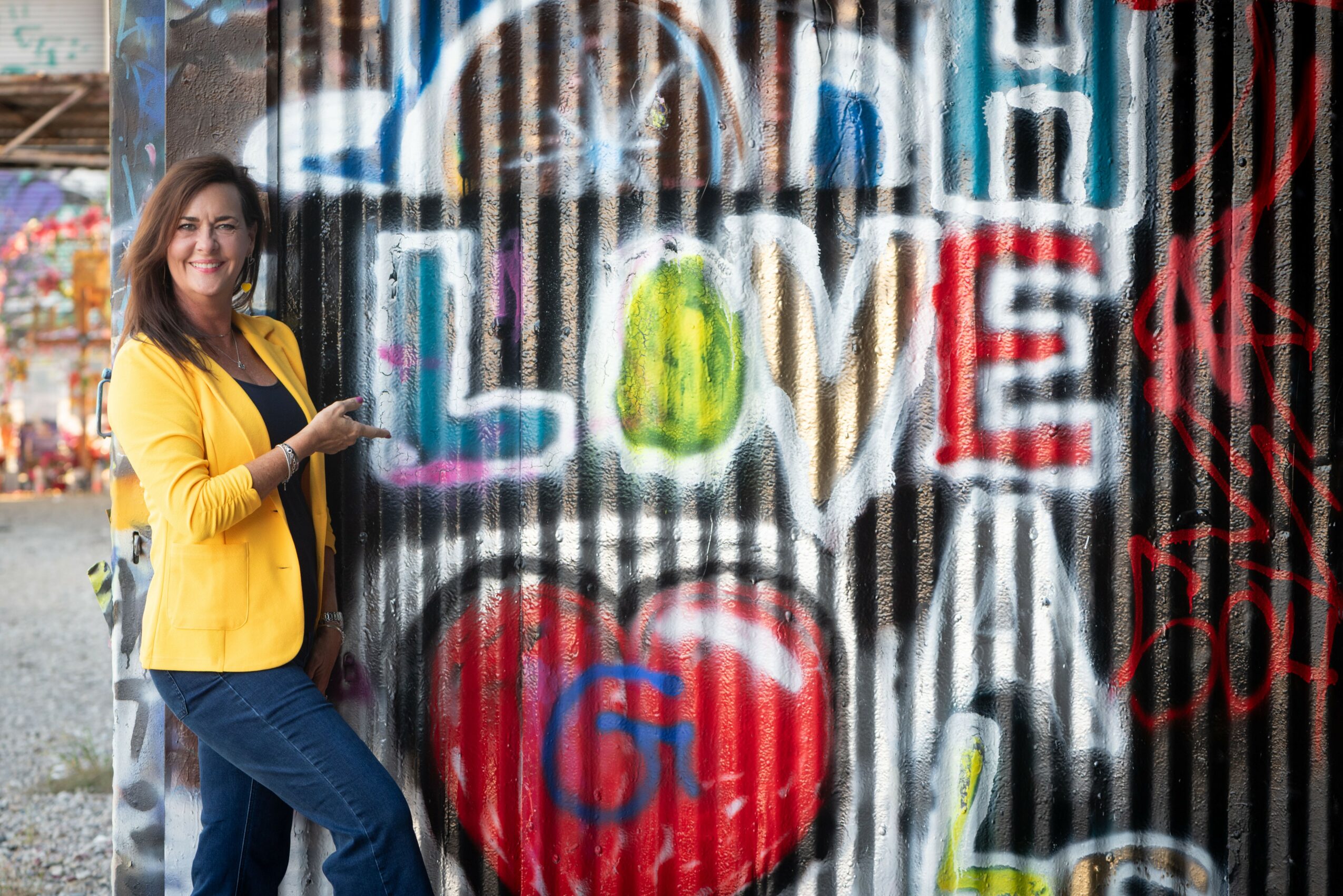 Woman in yellow jacket smiling next to graffiti with colorful words and a red heart, holding a phone.