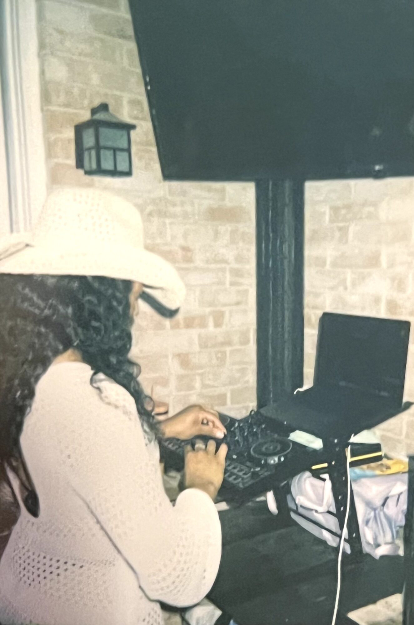 Person wearing a white hat and sweater using DJ equipment outdoors at night, with a brick wall and lamp in background.