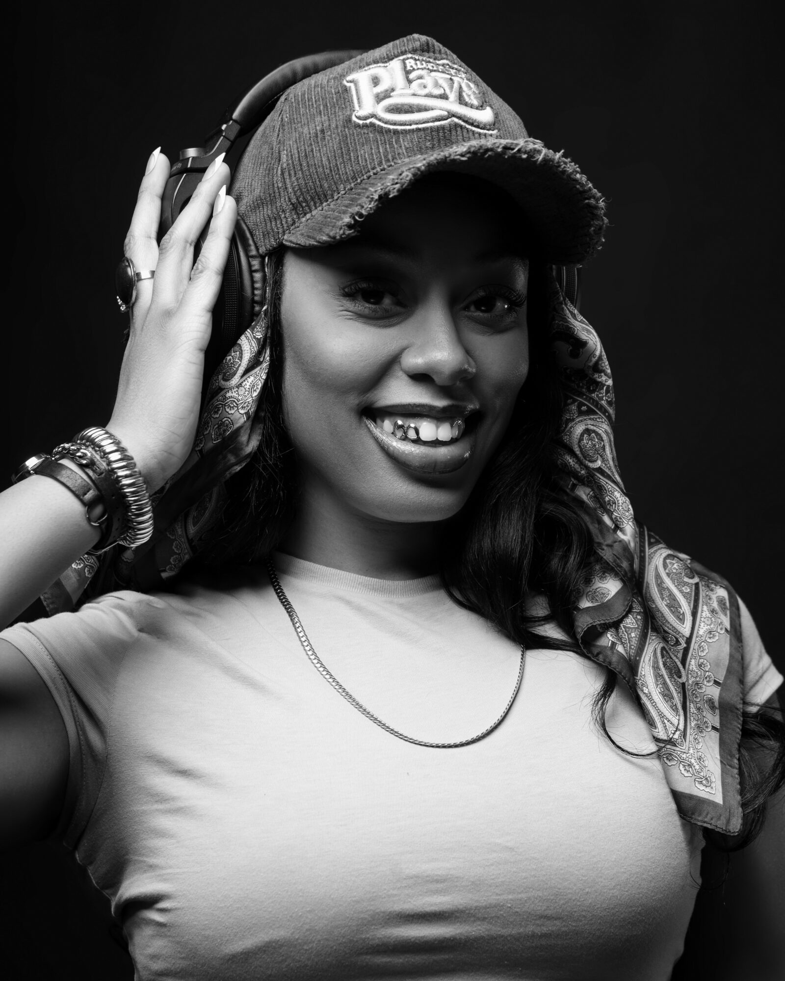 Woman wearing a baseball cap and headphones, smiling, with long hair and jewelry, in black and white.