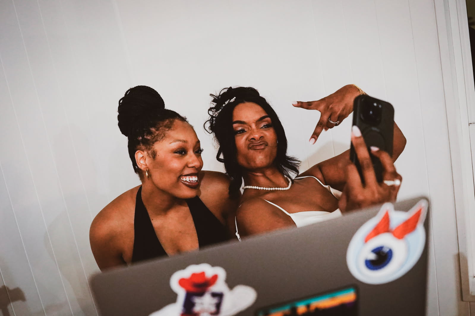 Two women taking a selfie together, smiling and making a peace sign, with a laptop in front of them.