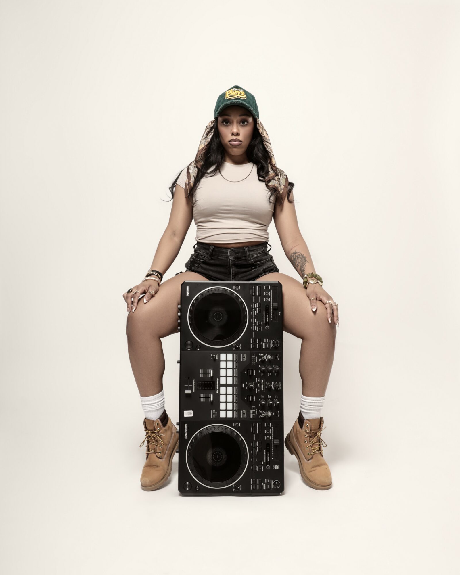 Young woman sitting on a DJ controller, wearing a cap, tank top, shorts, and boots, against a plain background.
