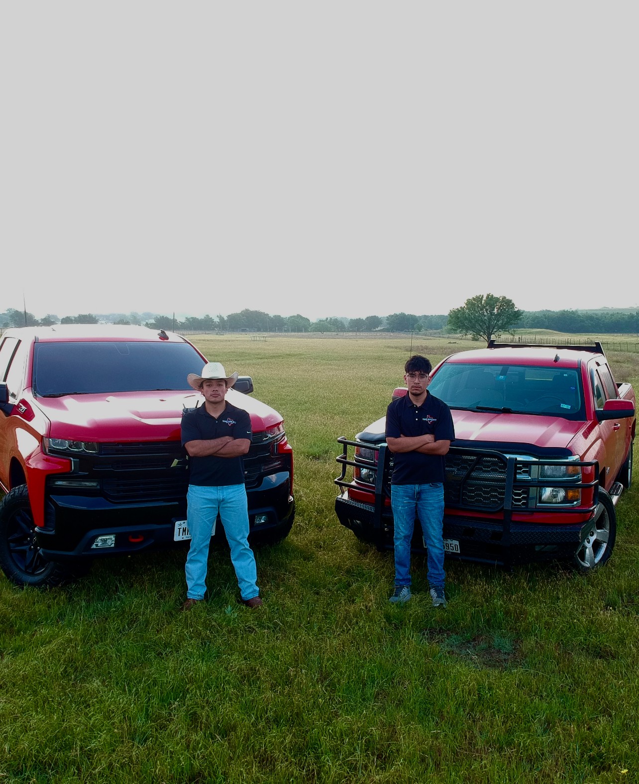 Two people stand in front of red vehicles on a grassy field, with arms crossed and looking at the camera.