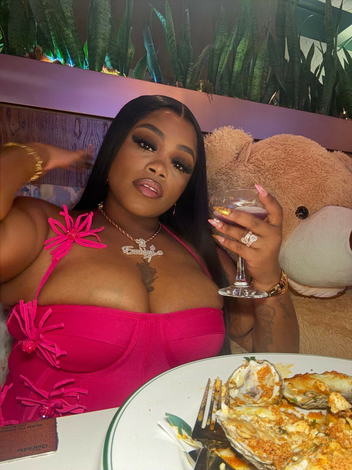 Woman in a pink dress with jewelry, sitting at a table with food, holding a fork, with a teddy bear in the background.