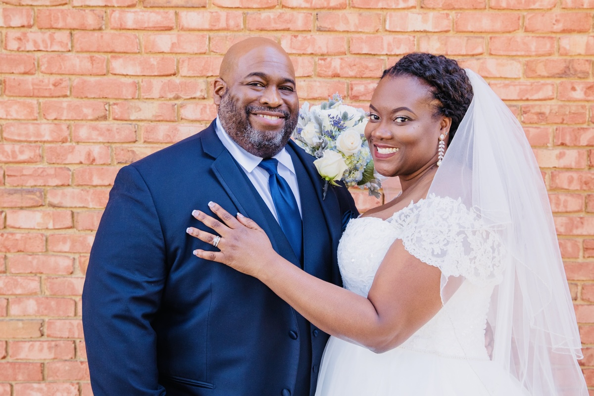 A bride and groom smiling, standing close together in front of a brick wall, with the bride holding a bouquet.