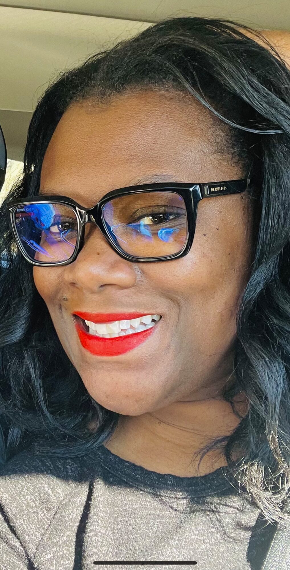 Woman with glasses smiling, wearing red lipstick, with dark wavy hair, in a car interior.