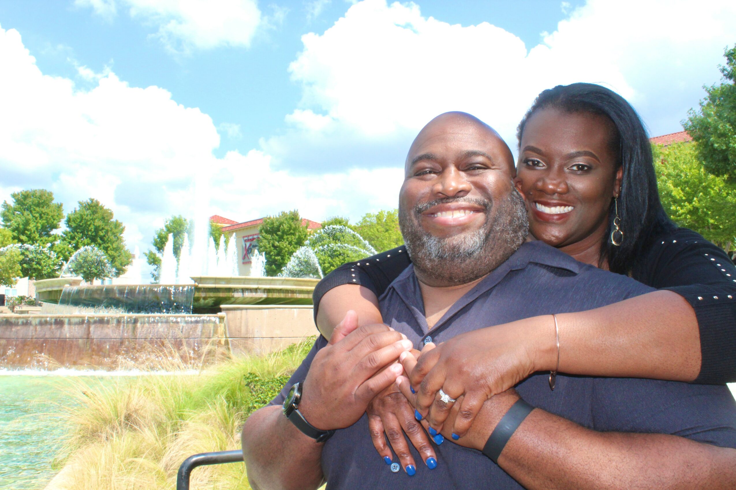 A smiling couple outdoors with a fountain and trees in the background, the woman hugging the man from behind.
