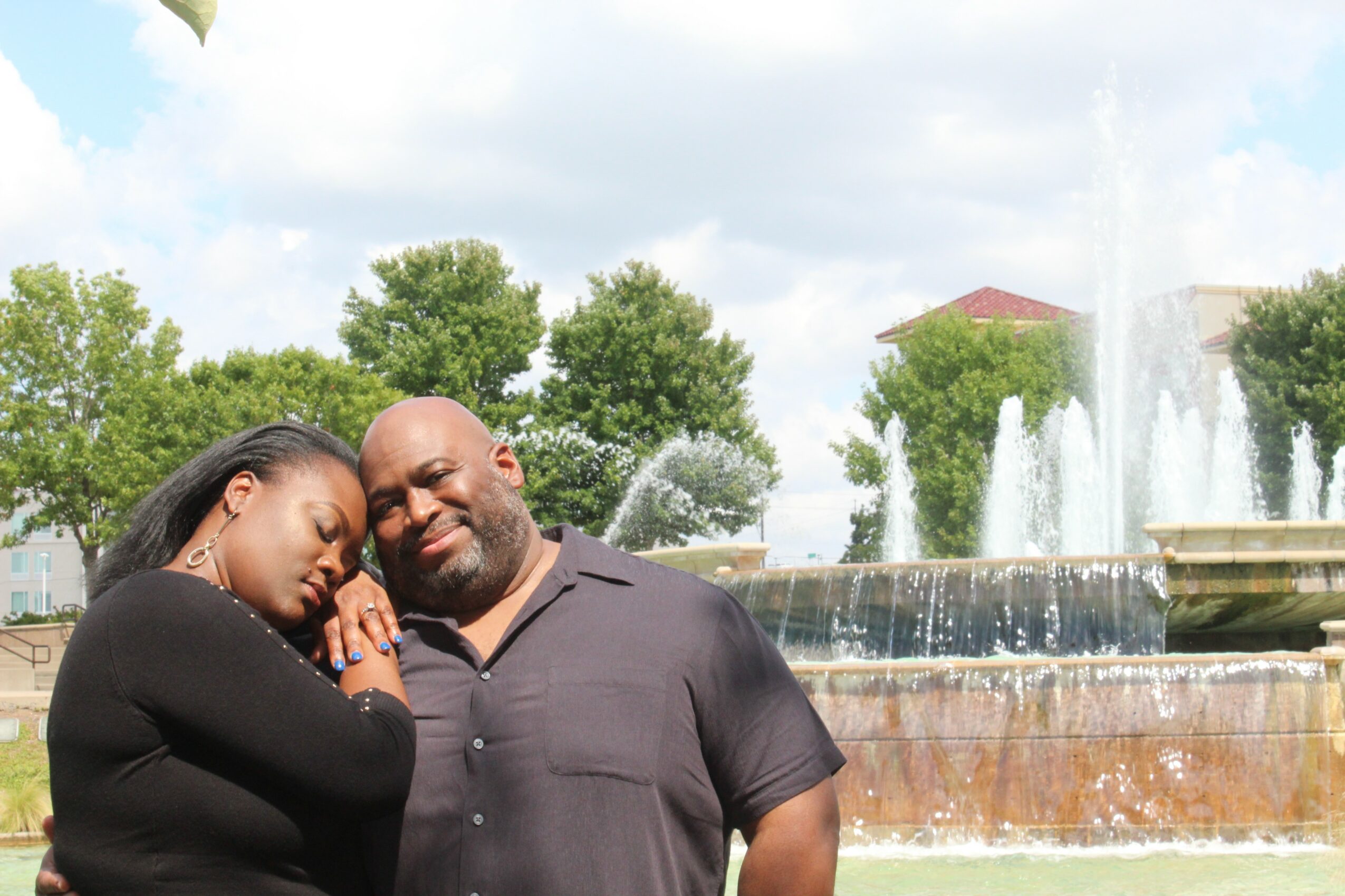 Two people smiling outdoors near a fountain with trees and a building in the background.