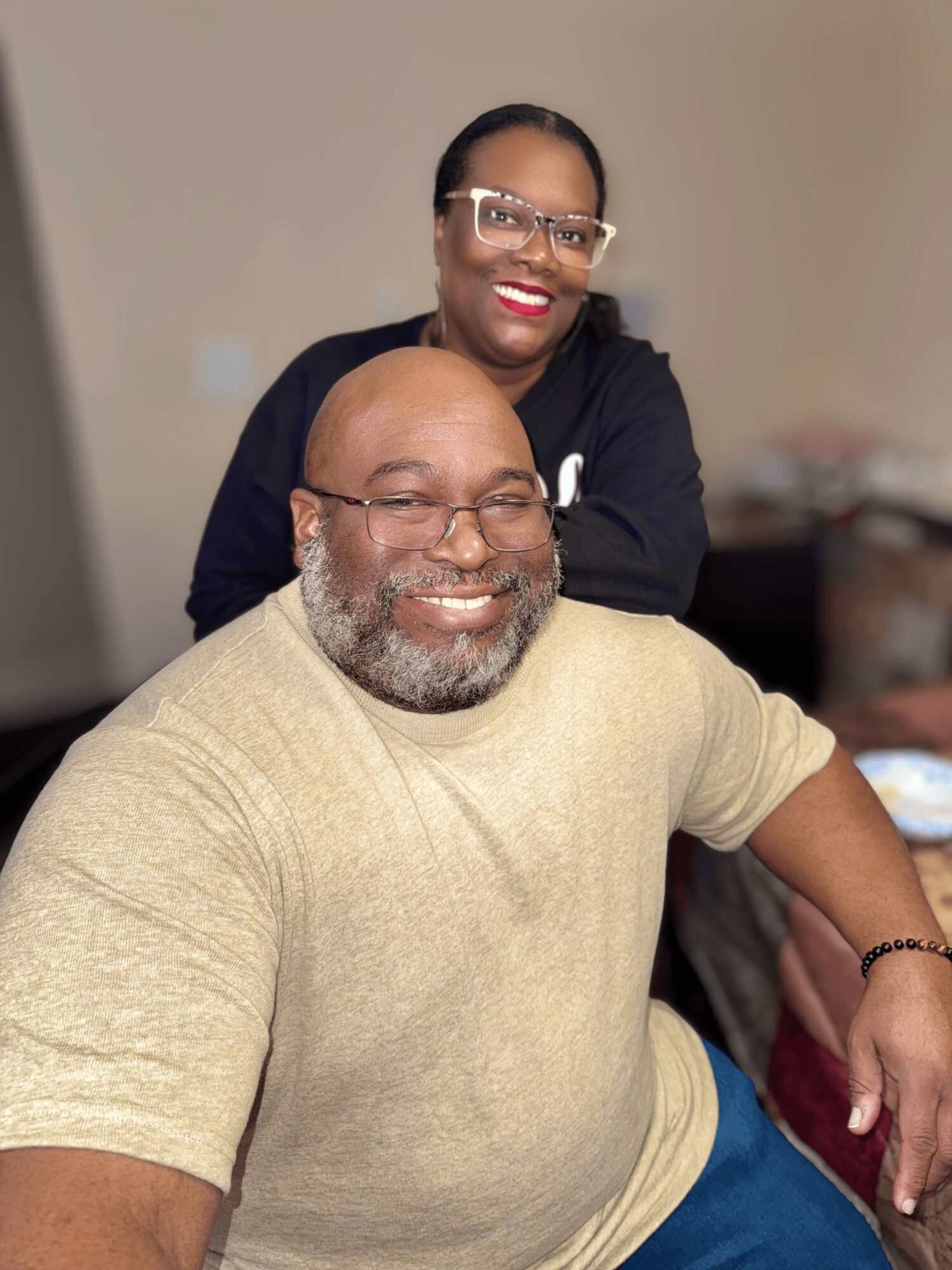 A smiling man with glasses and a beard, wearing a beige shirt, sitting indoors with a woman standing behind him.