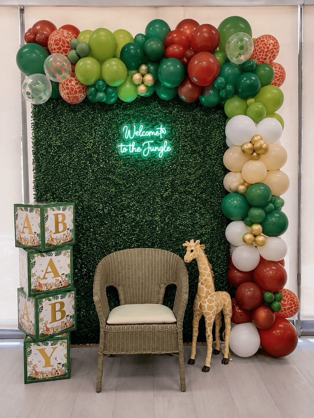 Balloon arch with green, red, and white balloons, a neon sign, a chair, a stuffed giraffe, and boxes with letters.