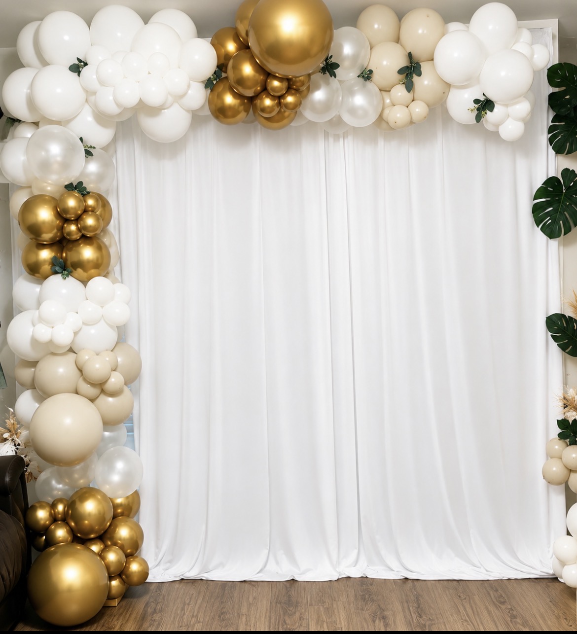 Decorative balloon arch with white, gold, and beige balloons over white curtains, with green leaves on the right side.