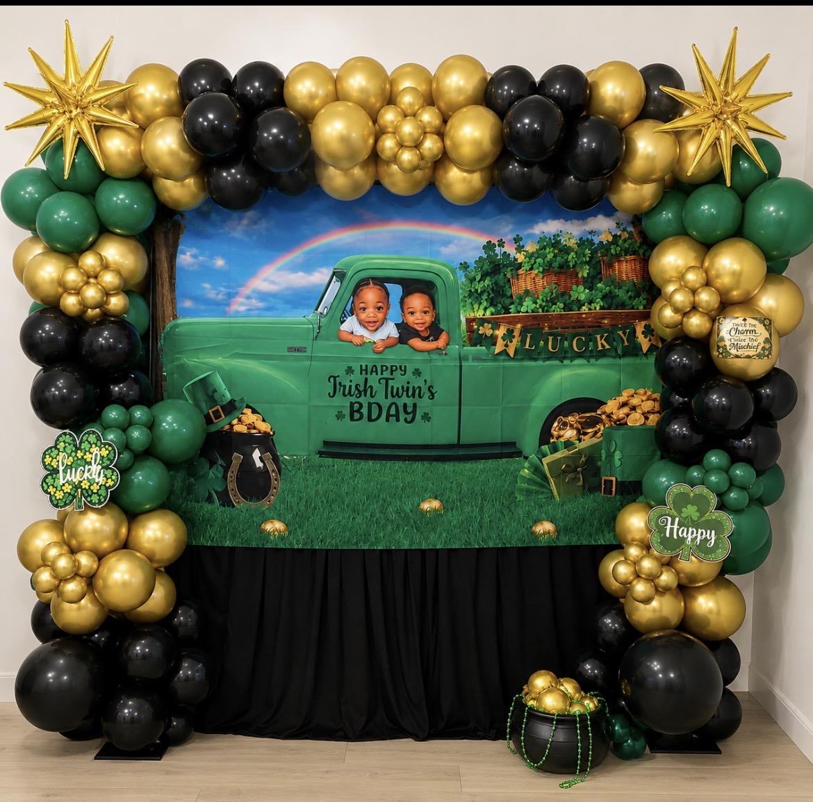 Decorative backdrop with children in a green truck, surrounded by black, gold, and green balloons, and festive decorations, for a birthday celebration.
