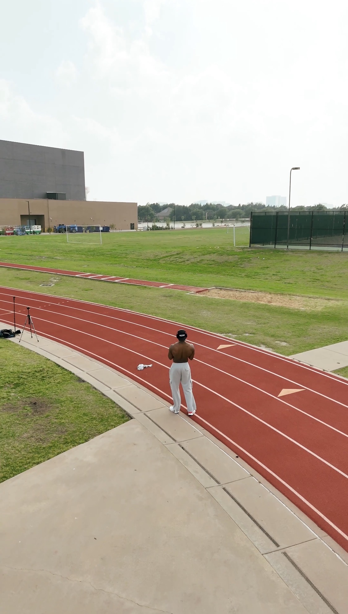 Person standing on a red running track near a grassy field with a building in the background.