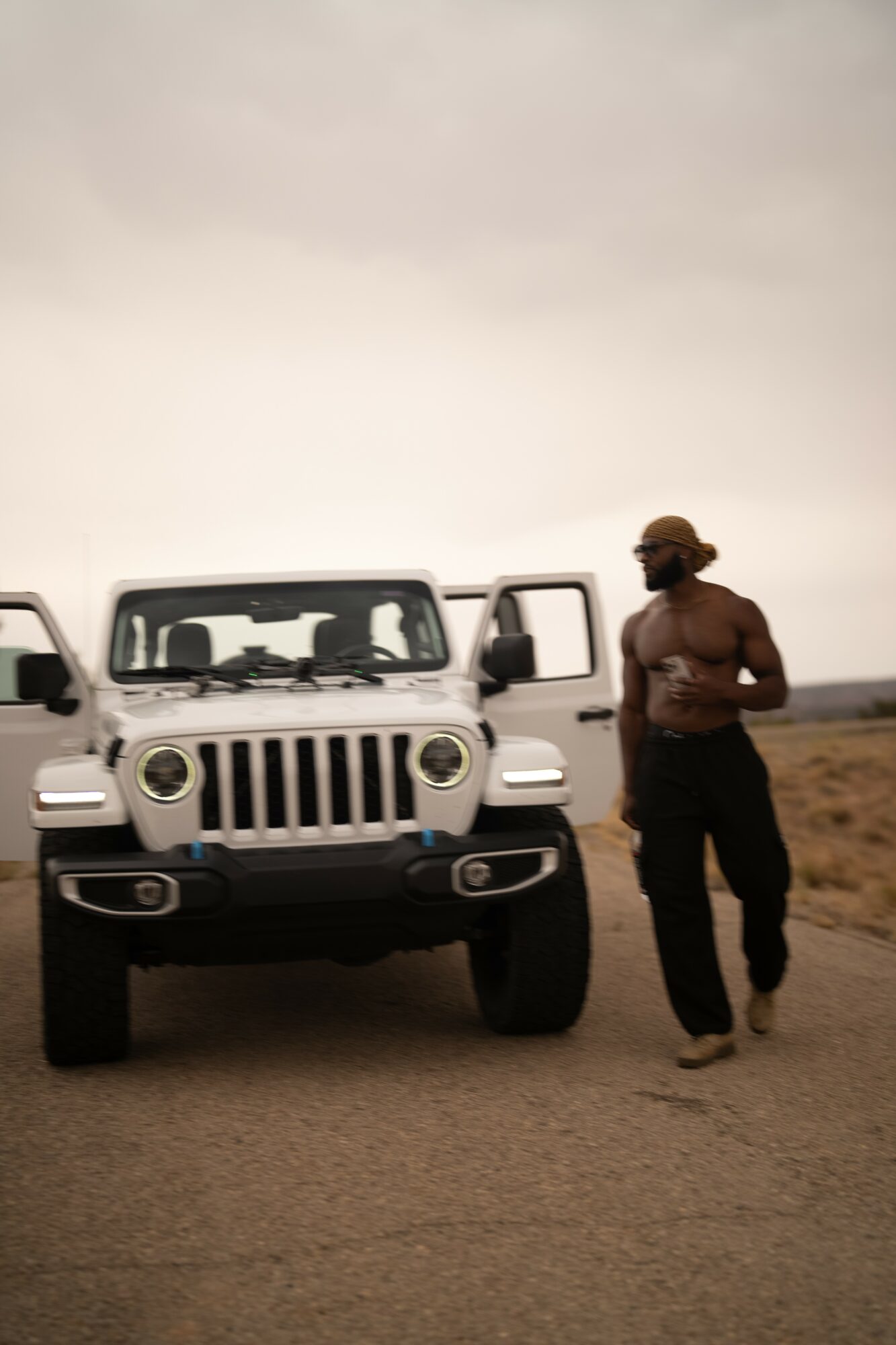 Person standing next to a white SUV on a dirt road, holding a phone, with open car doors, landscape background.