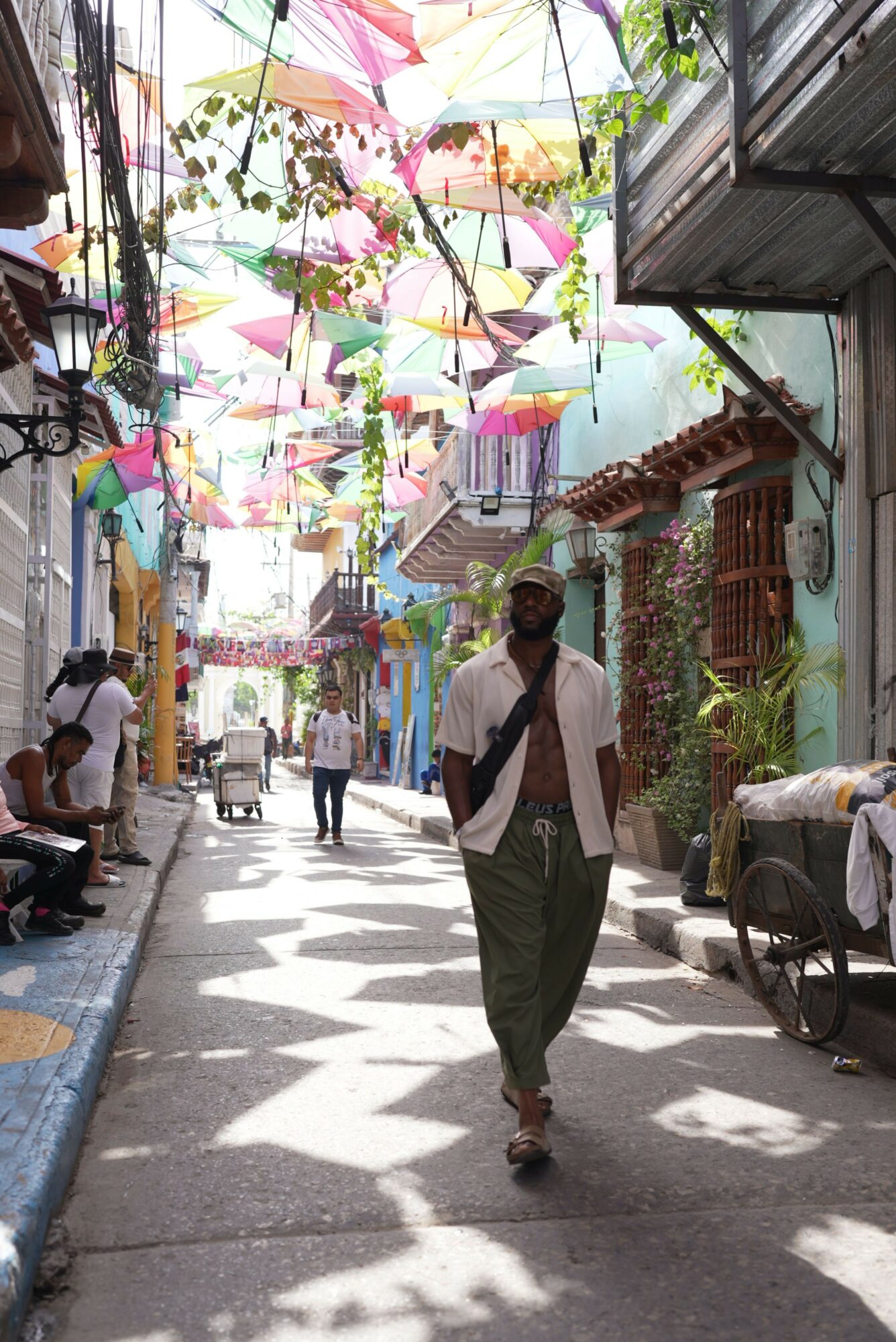 Street scene with people walking, colorful decorations, and shops, viewed from the side with shadows on the ground.