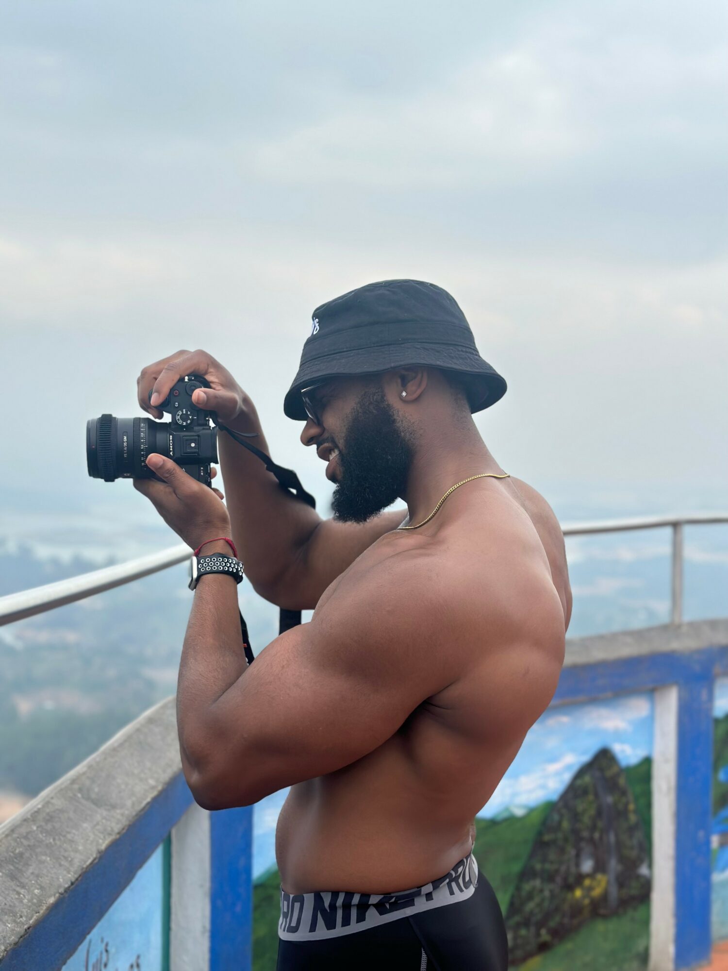 A shirtless man wearing a black bucket hat and sunglasses, holding a camera, standing outdoors near a railing with a cloudy sky.
