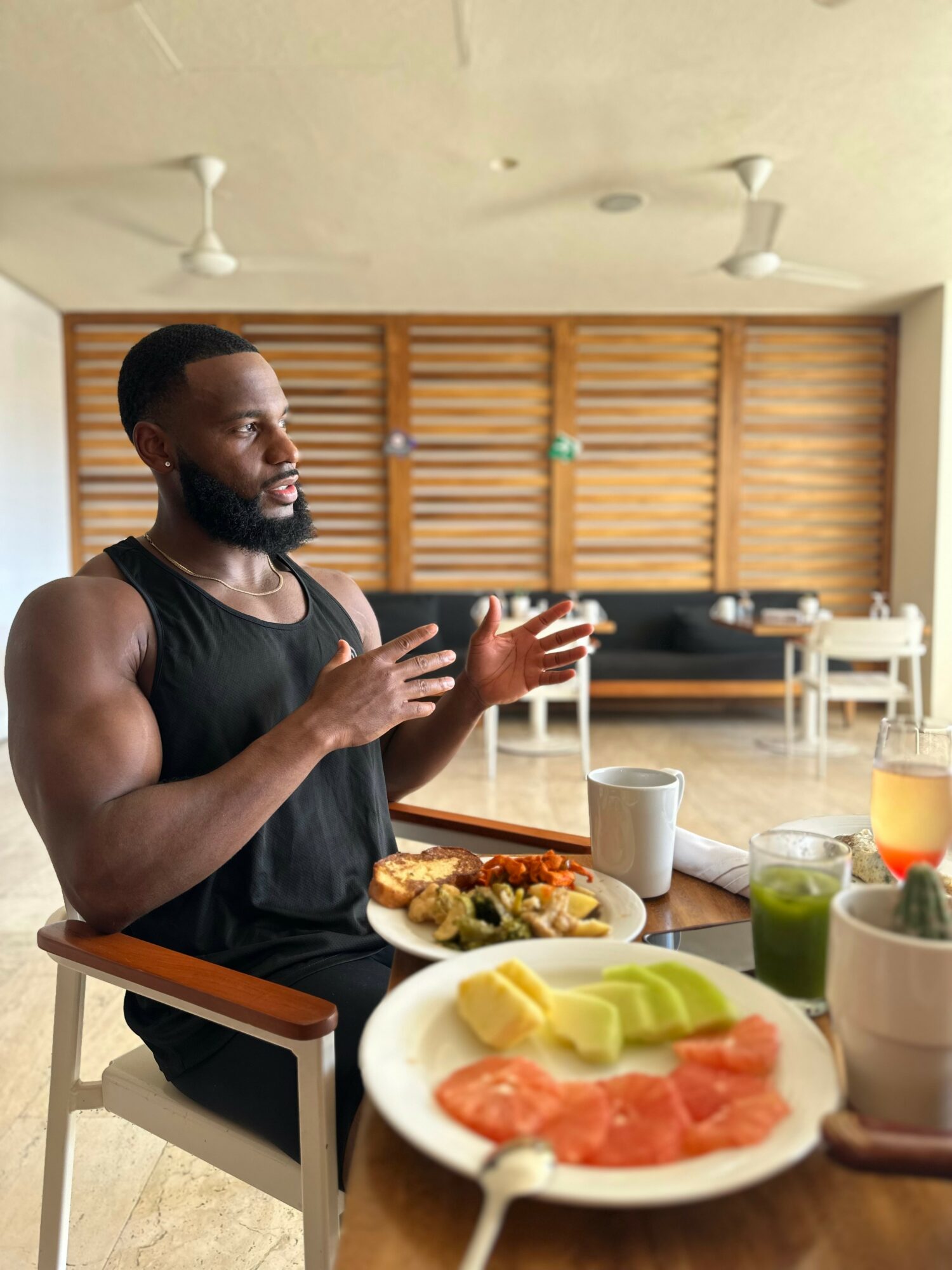 Man with a beard in a black sleeveless shirt sitting at a table with food and drinks, in a room with wooden wall panels.