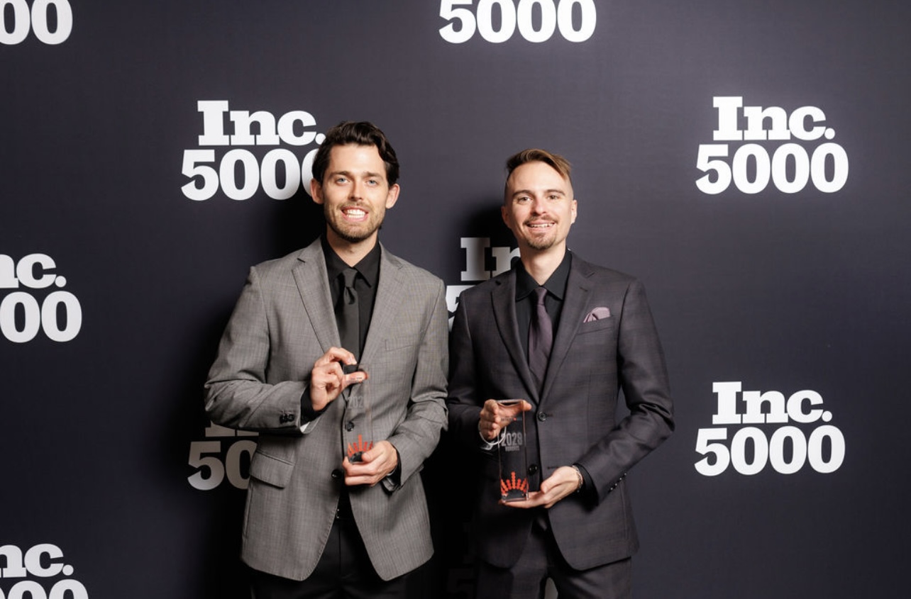Two men in suits holding awards, standing in front of a black backdrop with white text, smiling.