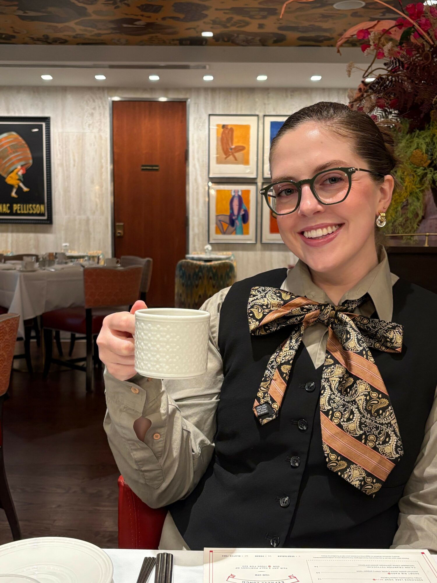 Woman with glasses smiling, holding a white mug, in an indoor setting with artwork and flowers in the background.