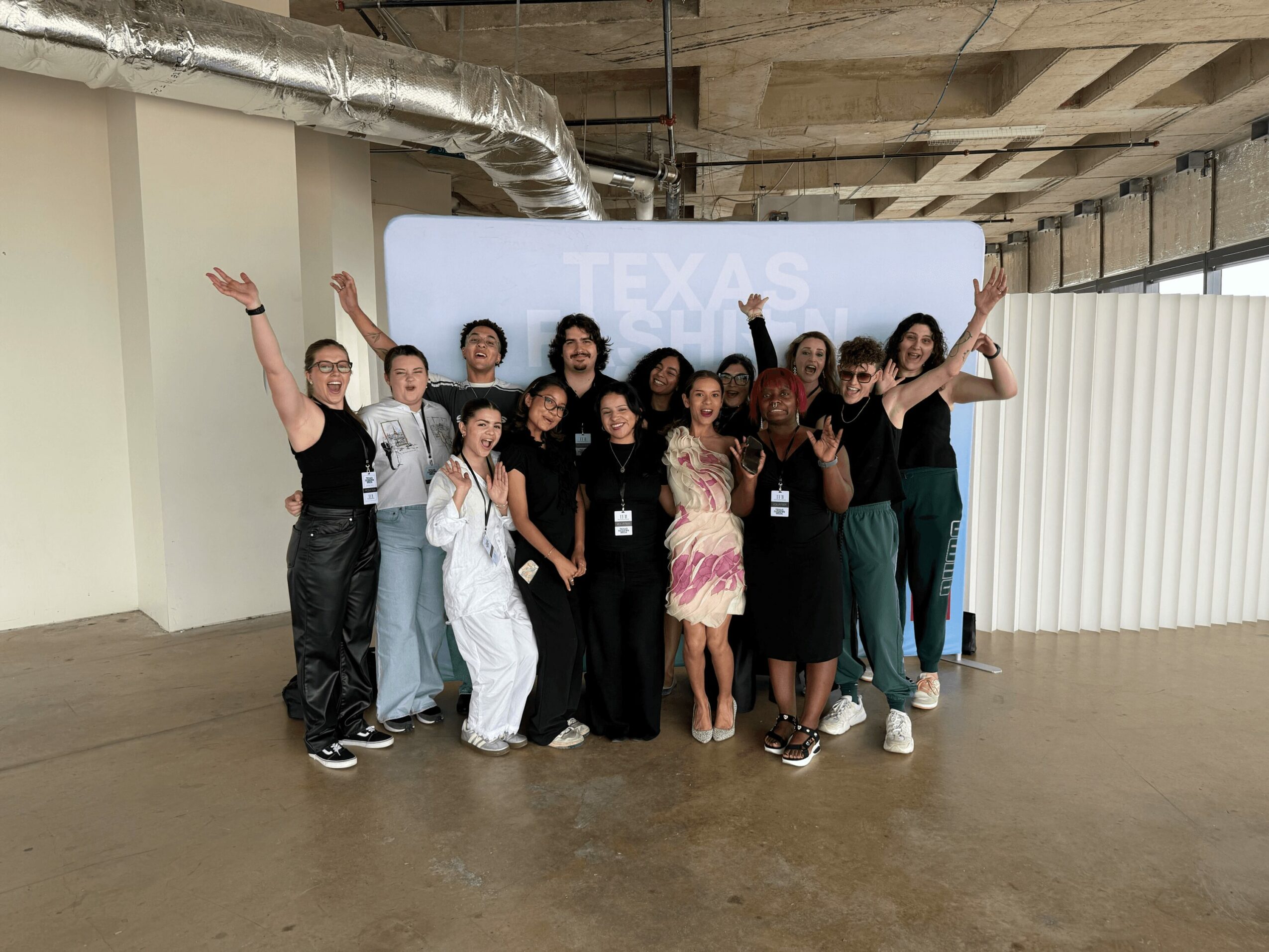 Group of people posing indoors in front of a white sign with partially visible text, some raising their arms, smiling.