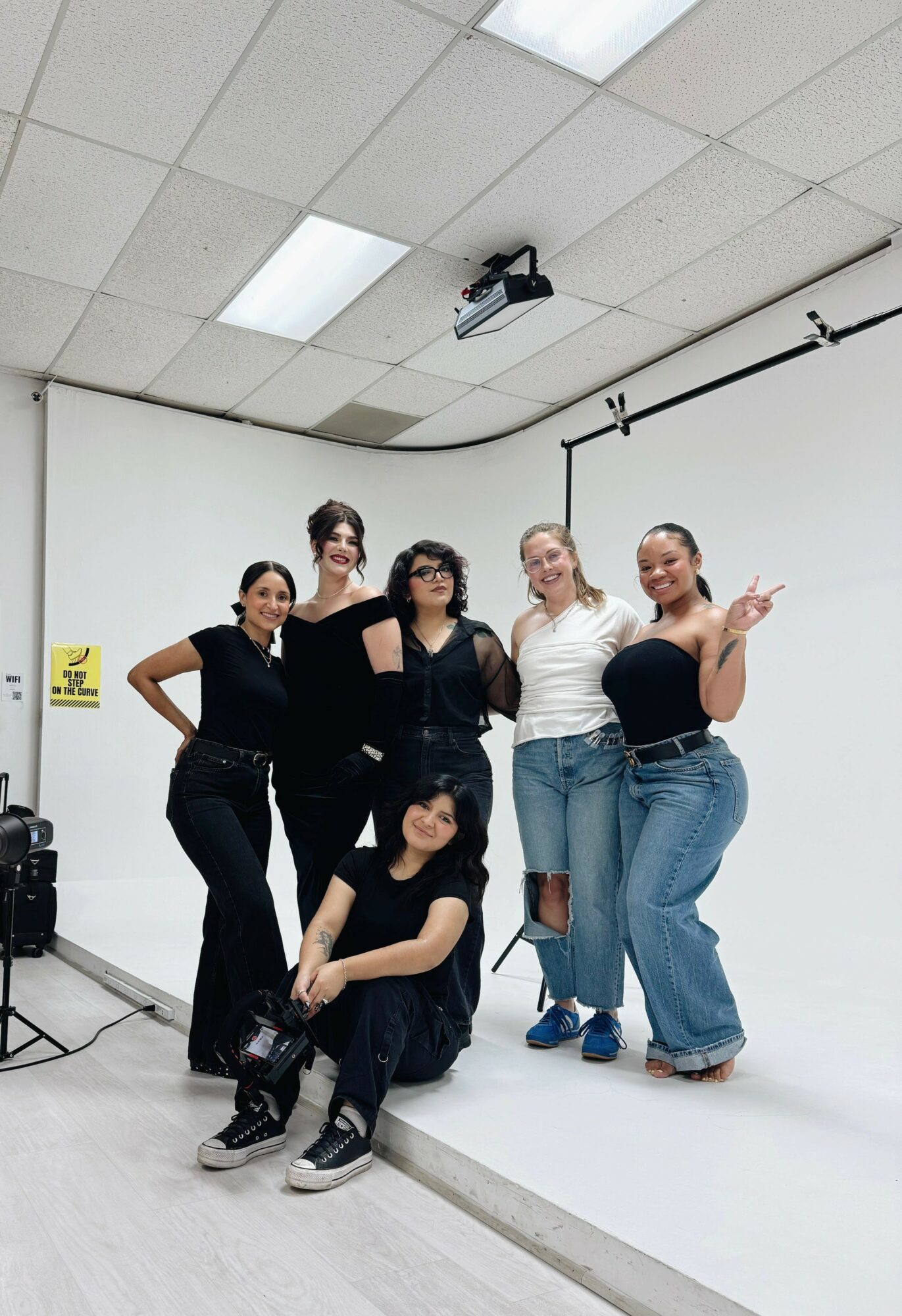 Six women pose together in a photography studio with a white backdrop and ceiling lights.