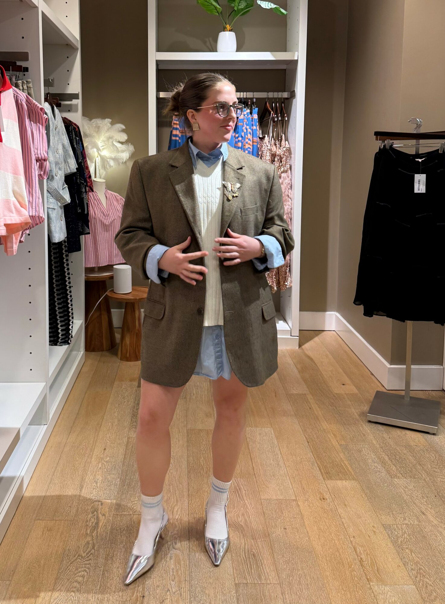 Person standing in clothing store, wearing oversized blazer, glasses, and patterned shoes, with clothing racks in background.