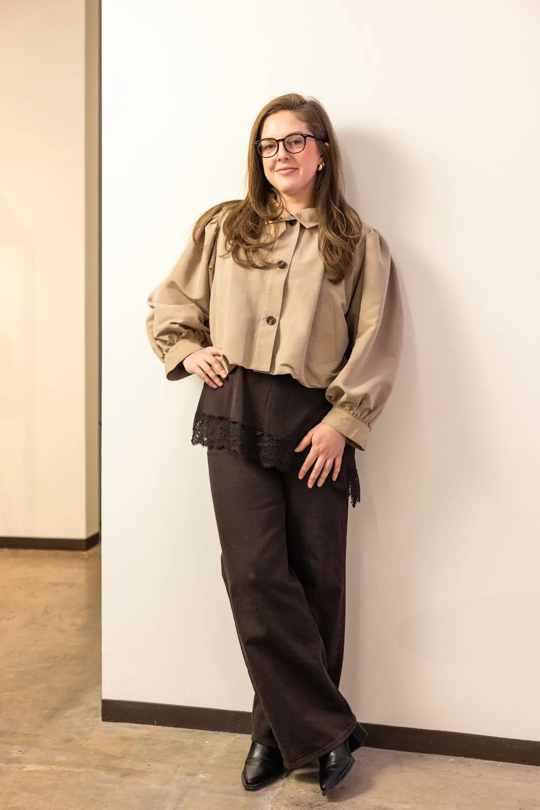 Young woman with glasses and long hair stands against a white wall, wearing a beige blouse, black pants, and black shoes.