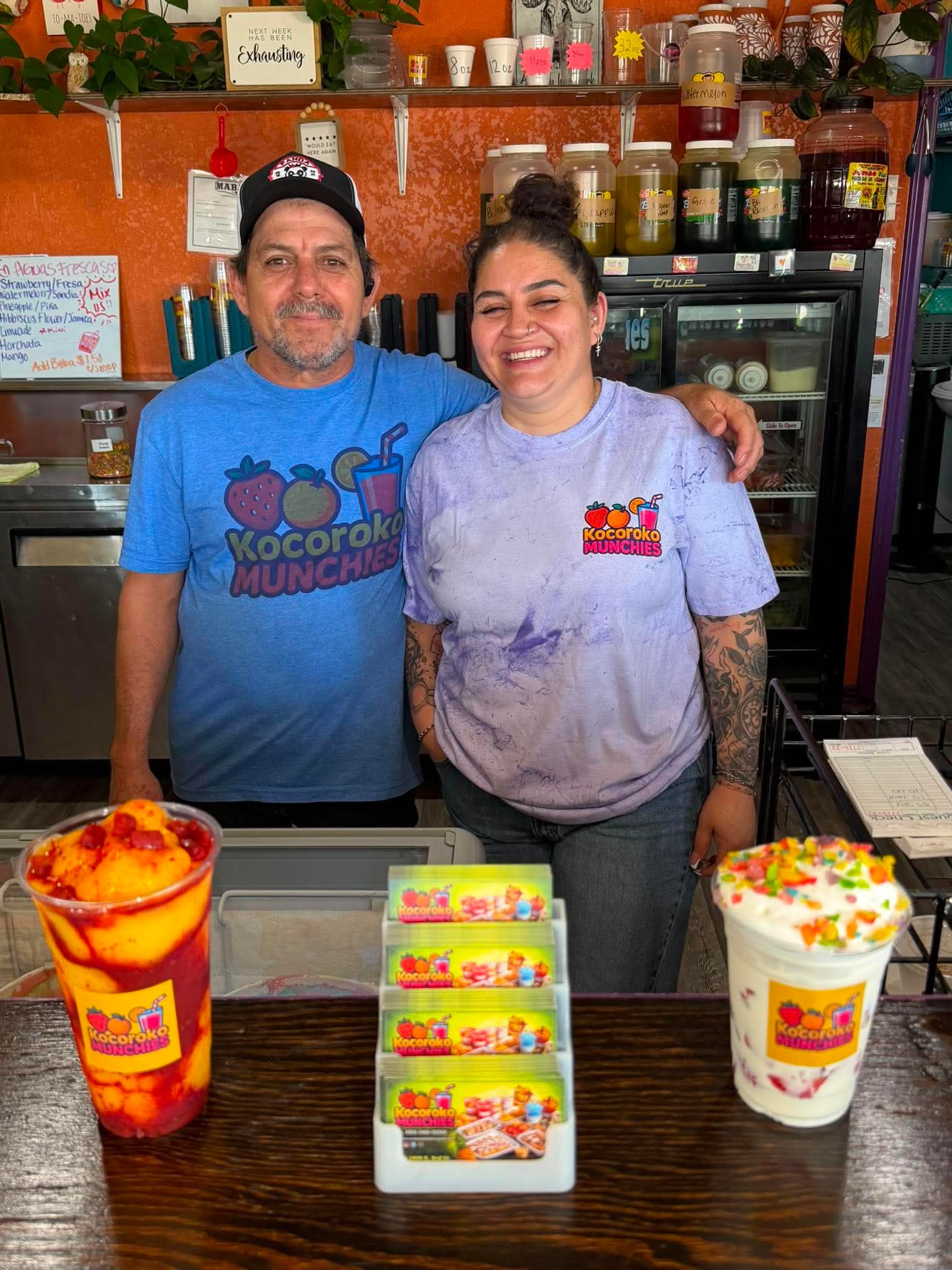 Two smiling people behind a counter with colorful drinks and candy, in a shop with shelves of jars and supplies.