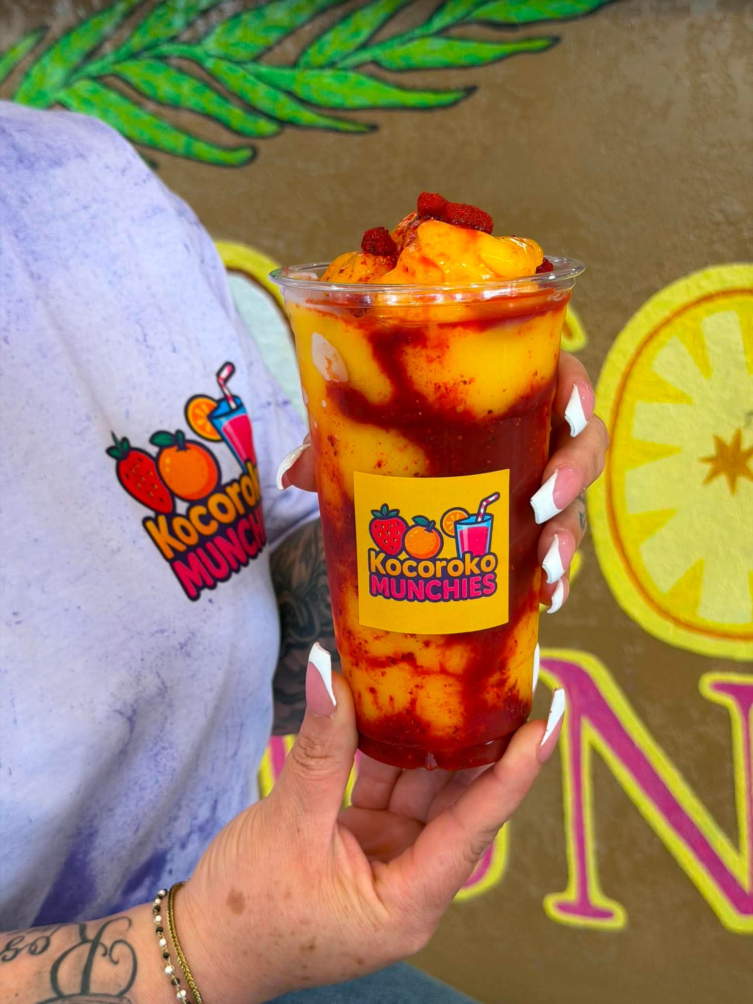 Person holding a tall glass of layered fruit drink with yellow, red, and orange colors, in front of a colorful background.